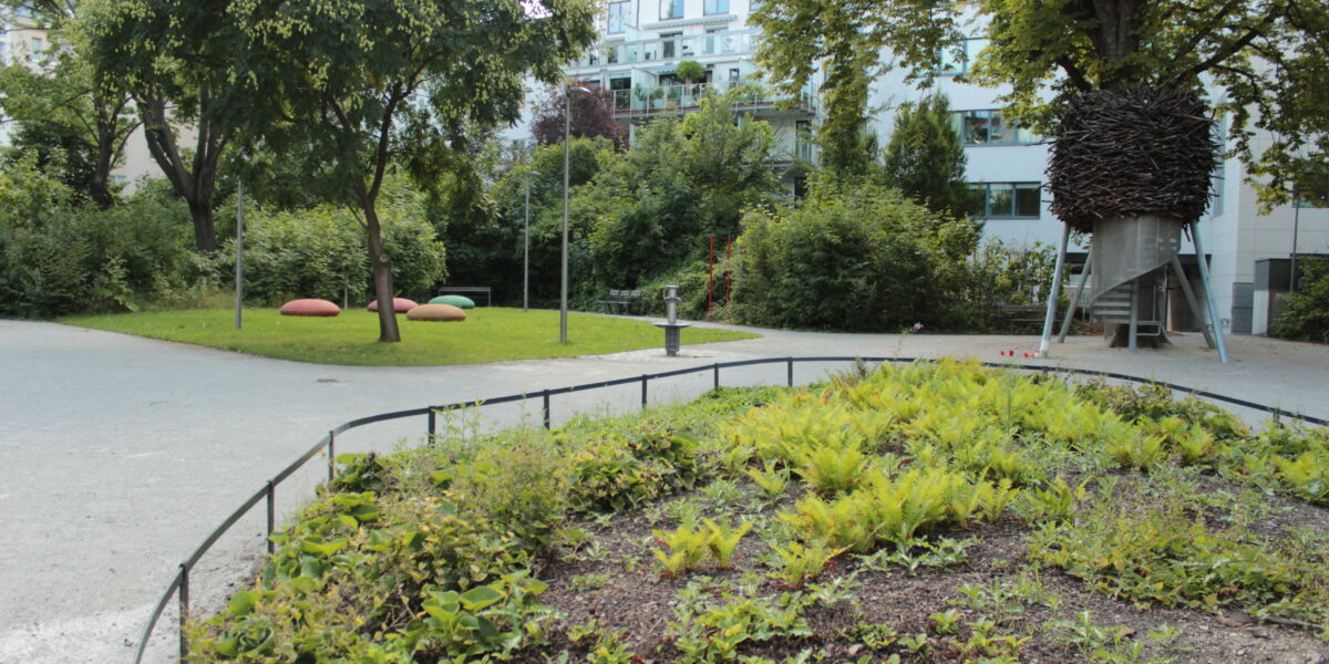 Ein kleiner Stadtpark mit grünen Sträuchern, Bäumen, einem gepflasterten Weg, großen Steinen und einer Metallkonstruktion mit dunklem, nestartigem Aussehen auf Stelzen. Im Hintergrund sind Wohnhäuser zu sehen.