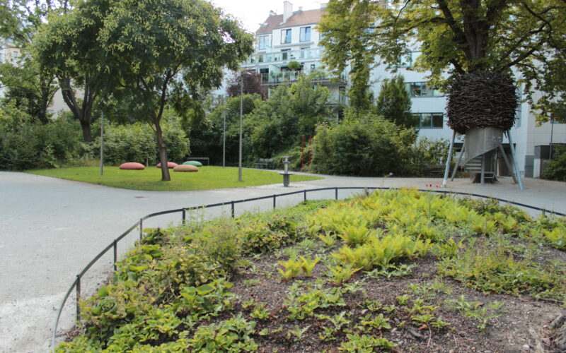 Ein kleiner Stadtpark mit grünen Sträuchern, Bäumen, einem gepflasterten Weg, großen Steinen und einer Metallkonstruktion mit dunklem, nestartigem Aussehen auf Stelzen. Im Hintergrund sind Wohnhäuser zu sehen.