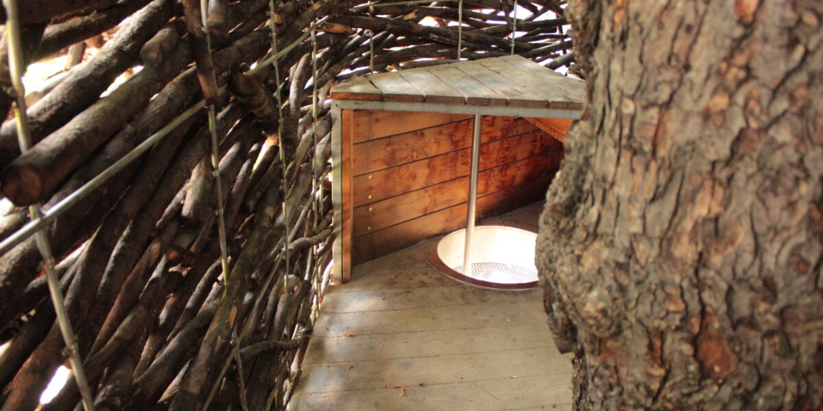Innenansicht eines rustikalen Baumhauses mit geflochtenen Astwänden, Holzboden und einem kleinen Holztisch über einer runden Öffnung.