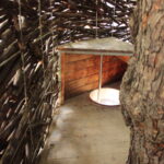 Innenansicht eines rustikalen Baumhauses mit geflochtenen Astwänden, Holzboden und einem kleinen Holztisch über einer runden Öffnung.