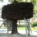 In einem Park im Freien wird ein großes künstliches Nest aus Stöcken um einen Baum herum gebaut und von Metallstangen gestützt.