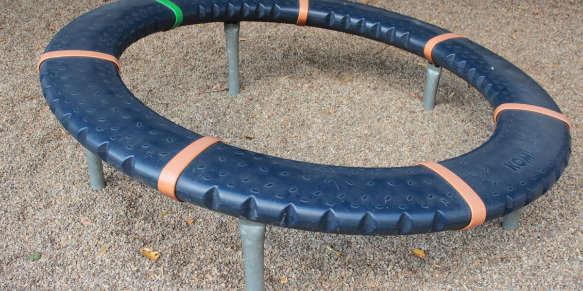 Ein kreisförmiger Spielplatz mit schwarzer, strukturierter Oberfläche, orangefarbenen Trennlinien und einem grünen Abschnitt, der von Metallfüßen getragen wird und auf einem Kiesboden steht.