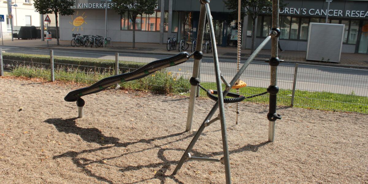 Ein Spielplatz mit einer schwarzen Wippe und einer runden Schaukel, umgeben von Kies, befindet sich in der Nähe einer Straße mit einem Gebäude und Bäumen im Hintergrund.
