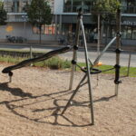 Ein Spielplatz mit einer schwarzen Wippe und einer runden Schaukel, umgeben von Kies, befindet sich in der Nähe einer Straße mit einem Gebäude und Bäumen im Hintergrund.