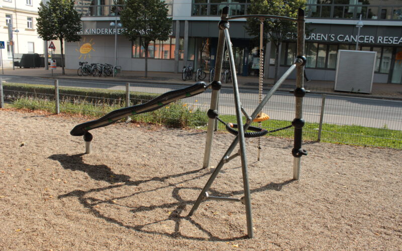Ein Spielplatz mit einer schwarzen Wippe und einer runden Schaukel, umgeben von Kies, befindet sich in der Nähe einer Straße mit einem Gebäude und Bäumen im Hintergrund.