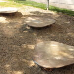 Drei flache, unregelmäßig geformte Metallplattformen mit Federn an der Unterseite befinden sich auf einer Kiesfläche in der Nähe eines Zauns, und auf den Plattformen sind einige Kreidezeichnungen zu sehen.