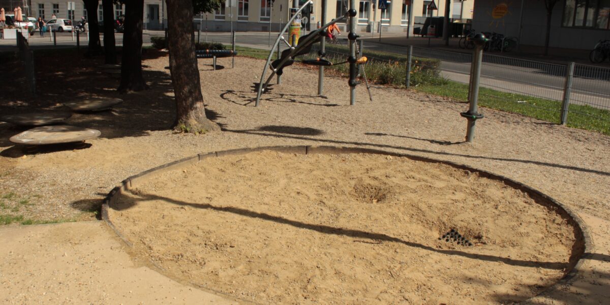 Ein kreisrunder Sandkasten mit aufgewühltem Sand befindet sich in einem schattigen Spielplatzbereich, im Hintergrund sind Klettergeräte und Gebäude zu sehen.