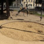 Ein kreisrunder Sandkasten mit aufgewühltem Sand befindet sich in einem schattigen Spielplatzbereich, im Hintergrund sind Klettergeräte und Gebäude zu sehen.