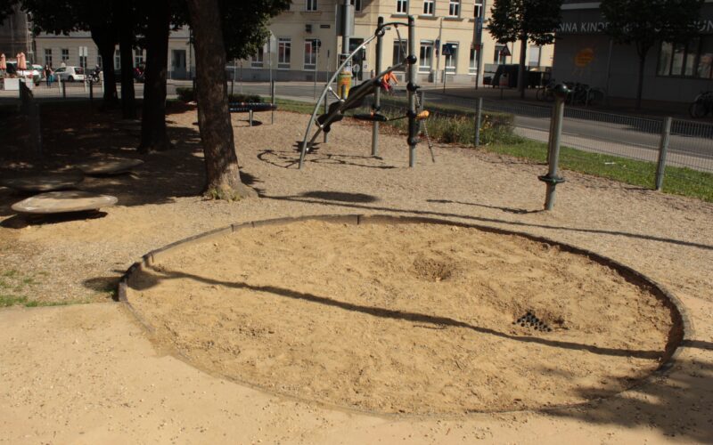 Ein kreisrunder Sandkasten mit aufgewühltem Sand befindet sich in einem schattigen Spielplatzbereich, im Hintergrund sind Klettergeräte und Gebäude zu sehen.