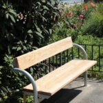 Eine Holzbank mit Armlehnen aus Metall steht auf einer gepflasterten Fläche neben grünen Büschen und blühenden Pflanzen in einem Garten.