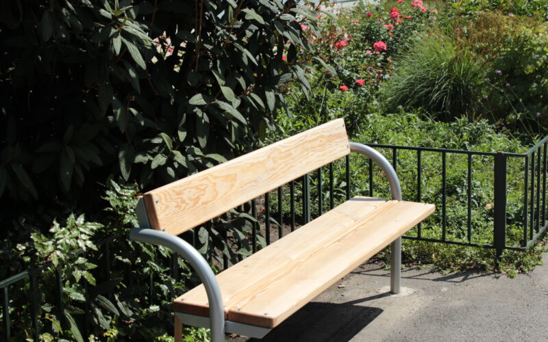 Eine Holzbank mit Armlehnen aus Metall steht auf einer gepflasterten Fläche neben grünen Büschen und blühenden Pflanzen in einem Garten.