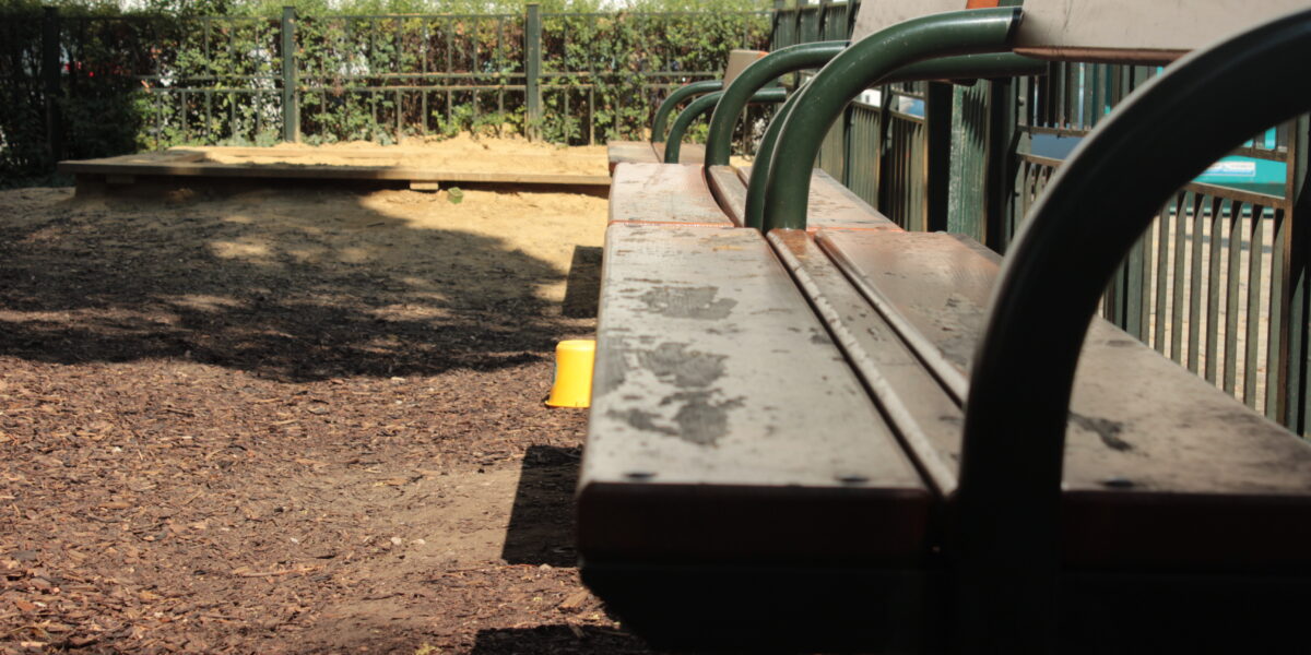 Eine hölzerne Parkbank mit grünen Metallarmlehnen vor einem eingezäunten Sandkastenbereich, mit Sonnenlicht und Schatten auf dem Boden.