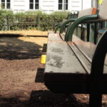 Eine hölzerne Parkbank mit grünen Metallarmlehnen vor einem eingezäunten Sandkastenbereich, mit Sonnenlicht und Schatten auf dem Boden.