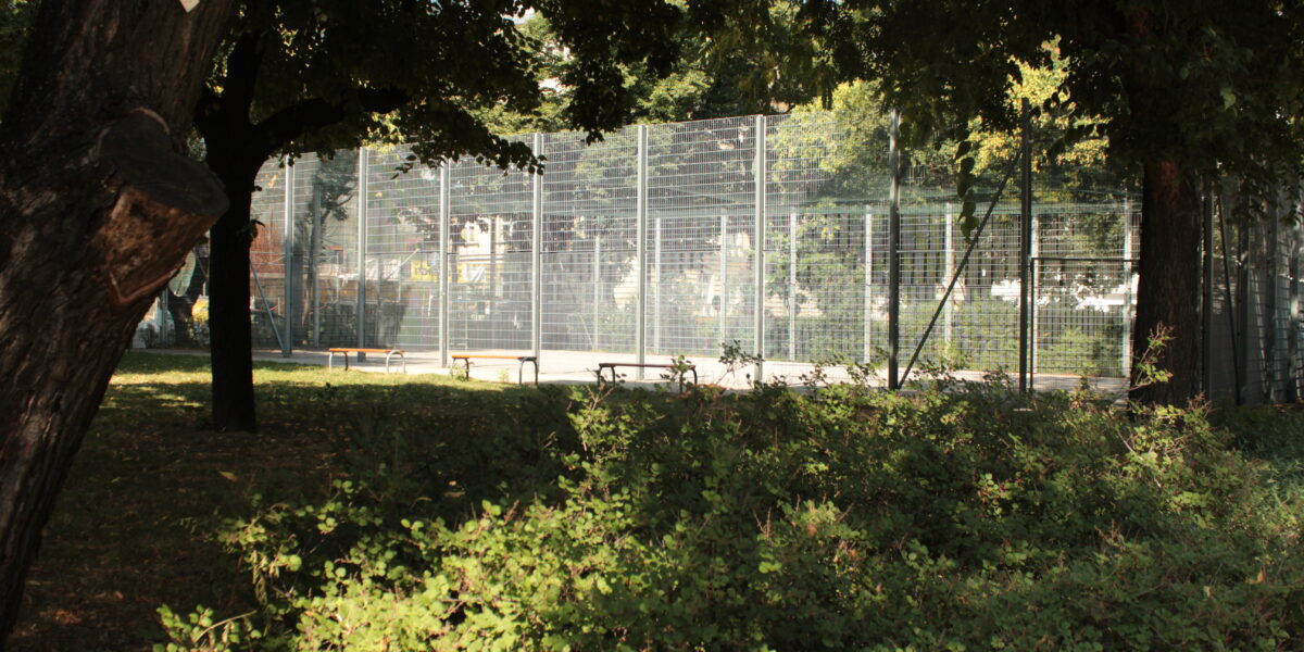 Ein eingezäunter Sportplatz ist von Bäumen und dichten Sträuchern umgeben, und an sonnigen Tagen kann man die Bänke am Rande sehen.