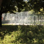 Ein eingezäunter Sportplatz ist von Bäumen und dichten Sträuchern umgeben, und an sonnigen Tagen kann man die Bänke am Rande sehen.