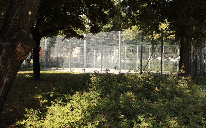 Ein eingezäunter Sportplatz ist von Bäumen und dichten Sträuchern umgeben, und an sonnigen Tagen kann man die Bänke am Rande sehen.