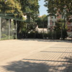 Ein leeres, umzäuntes Außengelände mit Drahtzaun und schattenspendenden Bäumen, begrenzt durch eine Backsteinmauer und ein Wohngebäude im Hintergrund.