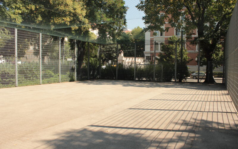 Ein leeres, umzäuntes Außengelände mit Drahtzaun und schattenspendenden Bäumen, begrenzt durch eine Backsteinmauer und ein Wohngebäude im Hintergrund.