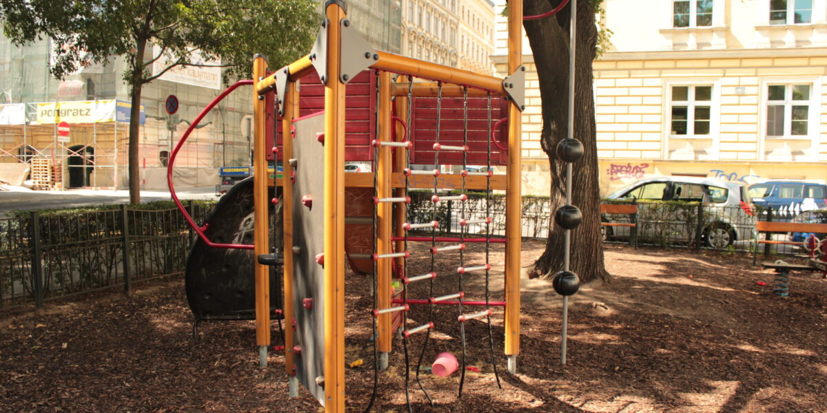 Ein Spielplatz-Klettergerüst mit Seilen, Leitern und Bällen, das auf Holzspänen unter Bäumen in der Nähe einer Stadtstraße und von Gebäuden steht.