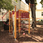 Ein Spielplatz-Klettergerüst mit Seilen, Leitern und Bällen, das auf Holzspänen unter Bäumen in der Nähe einer Stadtstraße und von Gebäuden steht.