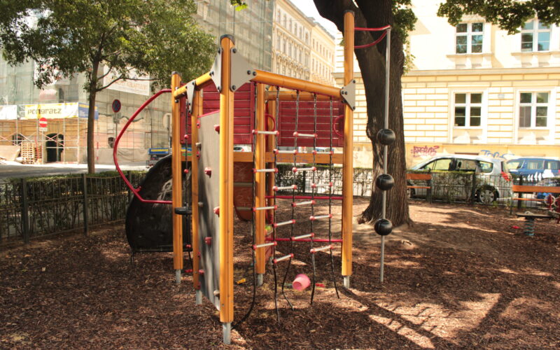 Ein Spielplatz-Klettergerüst mit Seilen, Leitern und Bällen, das auf Holzspänen unter Bäumen in der Nähe einer Stadtstraße und von Gebäuden steht.