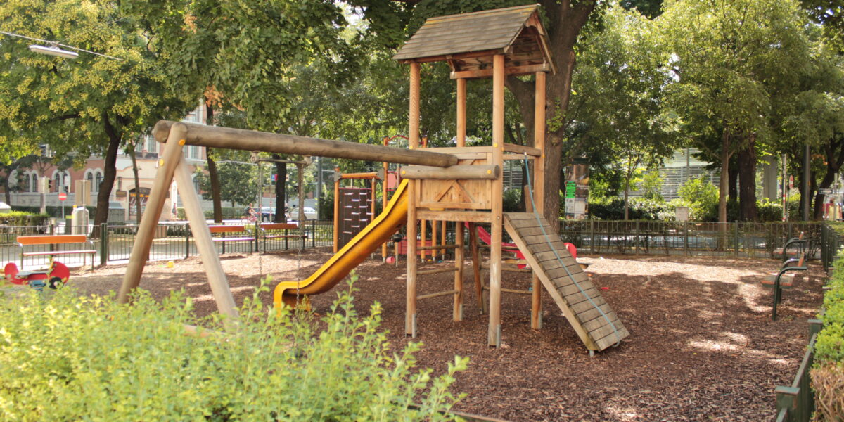 Spielplatz aus Holz mit einer gelben Rutsche, einer Kletterrampe und Schaukeln, umgeben von Bäumen und Büschen in einem Park im Freien.