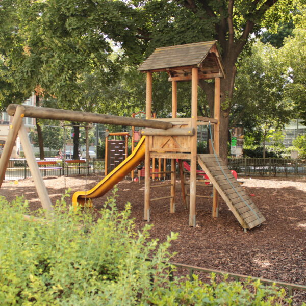 Spielplatz aus Holz mit einer gelben Rutsche, einer Kletterrampe und Schaukeln, umgeben von Bäumen und Büschen in einem Park im Freien.