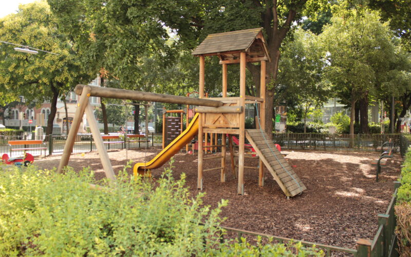 Spielplatz aus Holz mit einer gelben Rutsche, einer Kletterrampe und Schaukeln, umgeben von Bäumen und Büschen in einem Park im Freien.