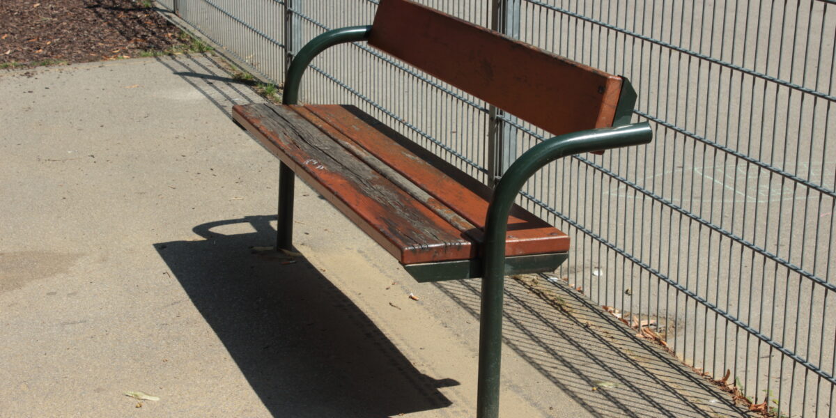 Eine abgenutzte Holzbank mit Metallbeinen und Rückenlehne steht auf einer Betonfläche neben einem Metallzaun in einem Außenbereich.