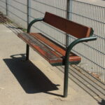 Eine abgenutzte Holzbank mit Metallbeinen und Rückenlehne steht auf einer Betonfläche neben einem Metallzaun in einem Außenbereich.