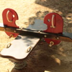 Eine Spielplatzwippe mit roten Sitzen in Form von Fischen steht auf sandigem Boden im Halbschatten.