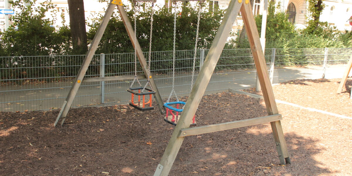 Holzschaukel mit zwei Schalensitzen auf einem mit Mulch bedeckten Spielplatz, umgeben von einem Metallzaun und Bäumen.