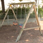 Holzschaukel mit zwei Schalensitzen auf einem mit Mulch bedeckten Spielplatz, umgeben von einem Metallzaun und Bäumen.