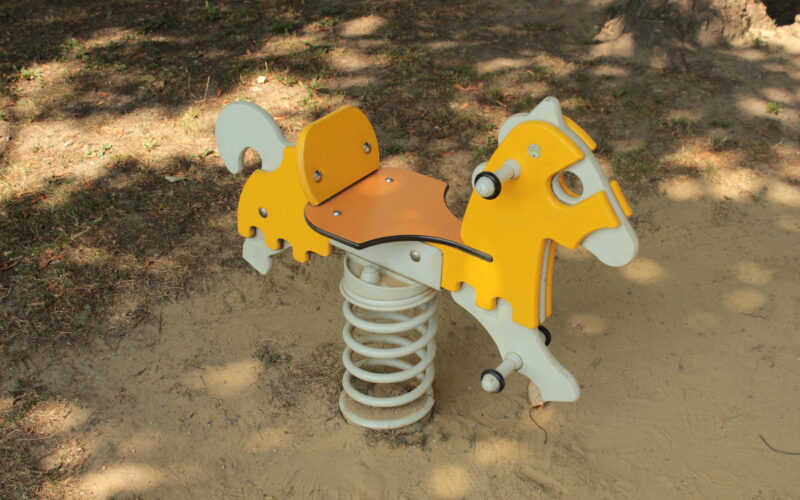 Ein gelbes und weißes gefedertes Schaukelpferd steht auf sandigem Boden im Halbschatten.