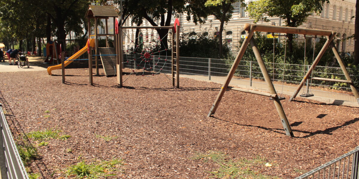 Leerer Spielplatz mit Schaukeln, einer Rutsche, Klettergeräten und Holzschnitzeldecke, umgeben von einem Metallzaun und Bäumen an einem sonnigen Tag.