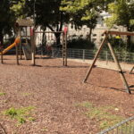 Leerer Spielplatz mit Schaukeln, einer Rutsche, Klettergeräten und Holzschnitzeldecke, umgeben von einem Metallzaun und Bäumen an einem sonnigen Tag.