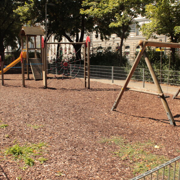 Leerer Spielplatz mit Schaukeln, einer Rutsche, Klettergeräten und Holzschnitzeldecke, umgeben von einem Metallzaun und Bäumen an einem sonnigen Tag.