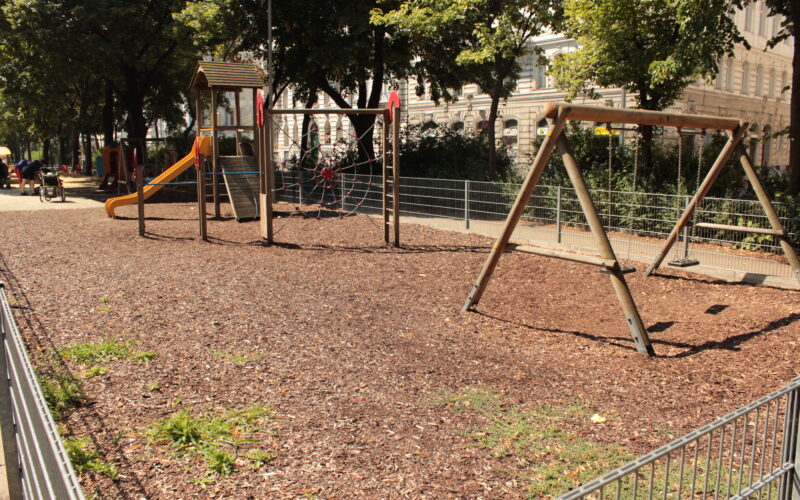Leerer Spielplatz mit Schaukeln, einer Rutsche, Klettergeräten und Holzschnitzeldecke, umgeben von einem Metallzaun und Bäumen an einem sonnigen Tag.