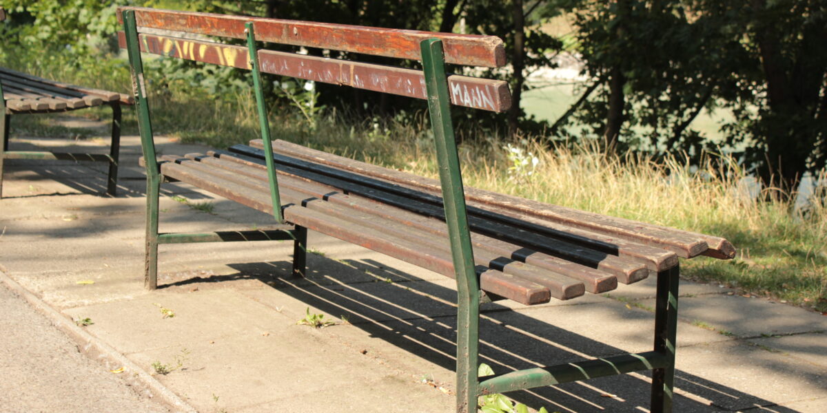 Eine abgenutzte Parkbank aus Holz mit Metallrahmen steht auf einem gepflasterten Weg neben Gras und Bäumen und wird vom Sonnenlicht beschattet.