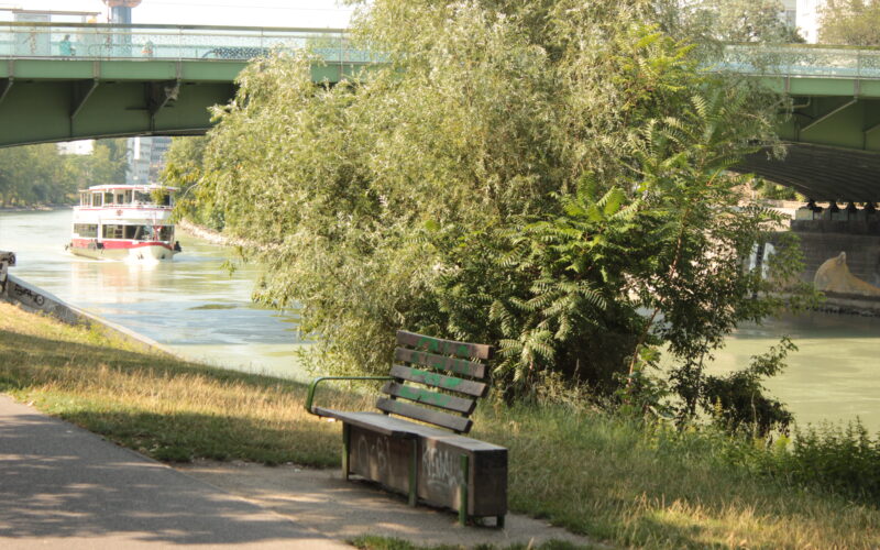 Eine grüne Bank steht an einem Uferweg, im Hintergrund eine Brücke und ein Boot auf dem Wasser. Bäume und Grün umgeben die Szene.