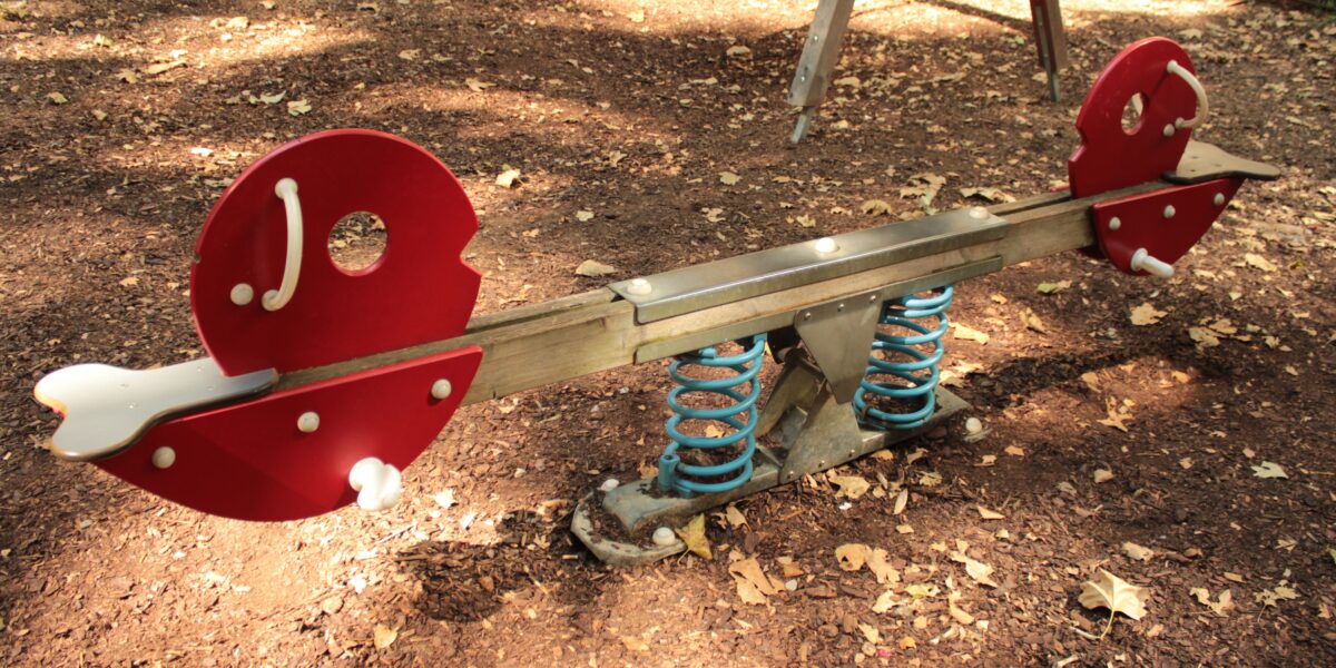 Auf einem Spielplatz steht eine rot-silberne Wippe mit blauen Federn auf einer mit trockenem Laub bedeckten Fläche.