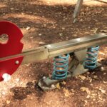 Auf einem Spielplatz steht eine rot-silberne Wippe mit blauen Federn auf einer mit trockenem Laub bedeckten Fläche.