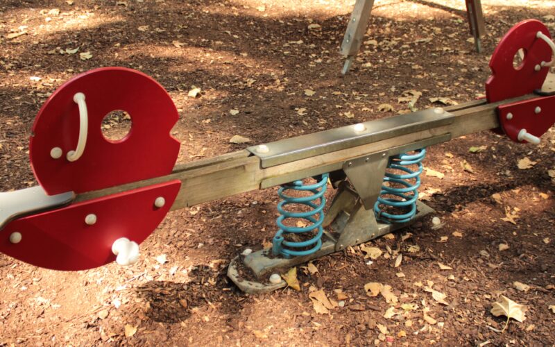 Auf einem Spielplatz steht eine rot-silberne Wippe mit blauen Federn auf einer mit trockenem Laub bedeckten Fläche.