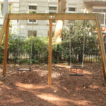 Eine hölzerne Schaukel mit zwei leeren Schaukeln steht auf einem unbefestigten Boden in einem kleinen Stadtpark, umgeben von einem Metallzaun und Grünpflanzen, mit einem Gebäude im Hintergrund.