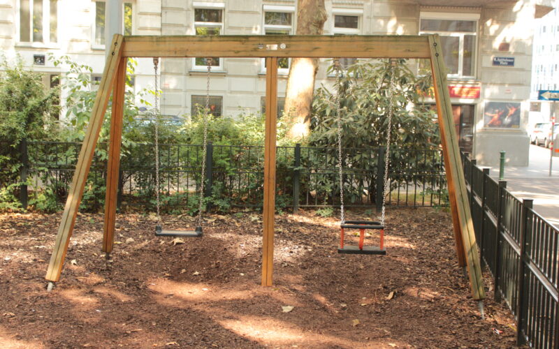 Eine hölzerne Schaukel mit zwei leeren Schaukeln steht auf einem unbefestigten Boden in einem kleinen Stadtpark, umgeben von einem Metallzaun und Grünpflanzen, mit einem Gebäude im Hintergrund.