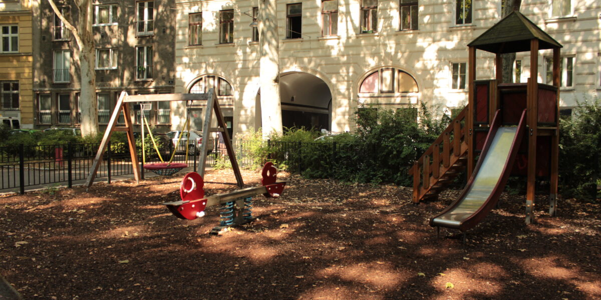 Ein Spielplatz mit einer Rutsche, Schaukeln und einer Wippe, umgeben von Wohnhäusern und Bäumen, im Sonnenlicht.