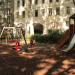 Ein Spielplatz mit einer Rutsche, Schaukeln und einer Wippe, umgeben von Wohnhäusern und Bäumen, im Sonnenlicht.