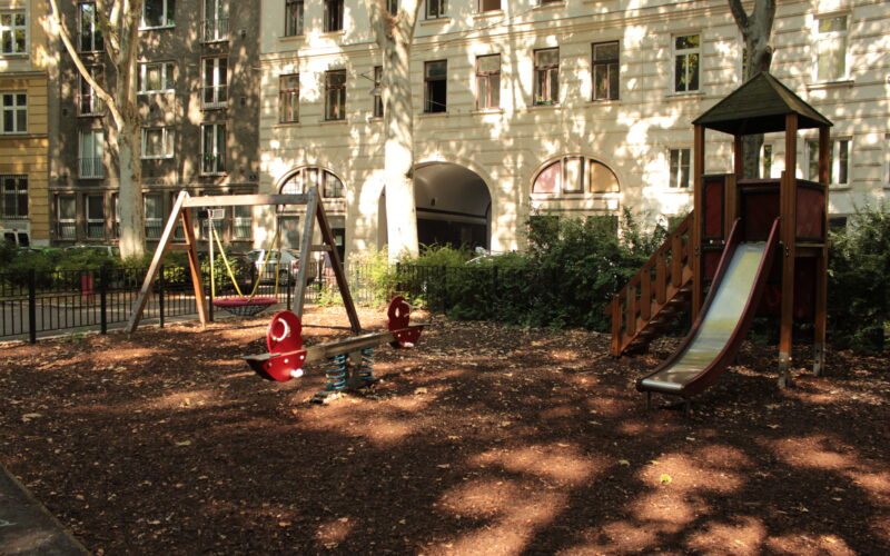 Ein Spielplatz mit einer Rutsche, Schaukeln und einer Wippe, umgeben von Wohnhäusern und Bäumen, im Sonnenlicht.