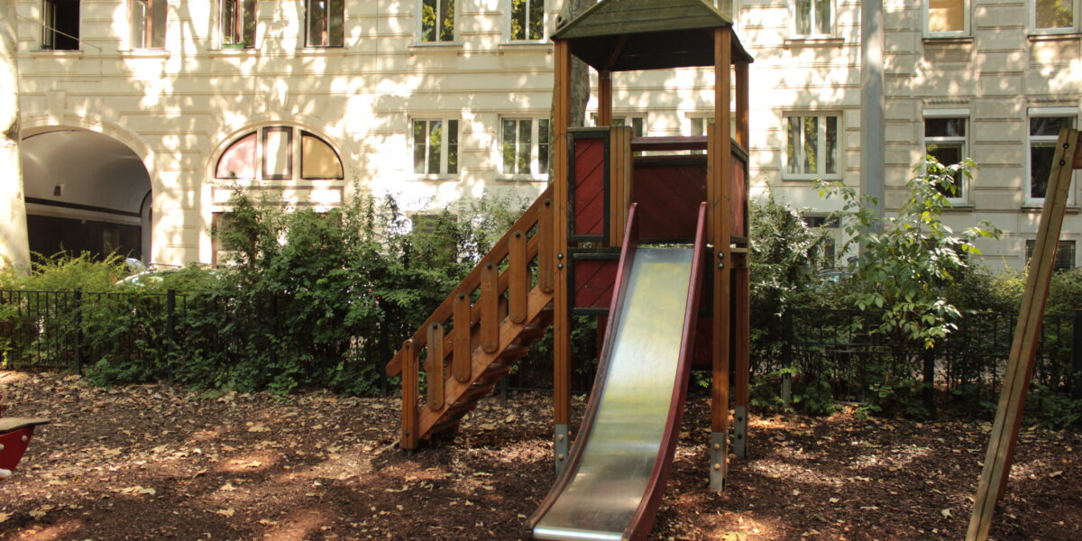 Eine Metallrutsche mit Holztreppe und Unterstand steht in einem schattigen Spielplatzbereich, umgeben von Bäumen und einem weißen Wohnhaus im Hintergrund.