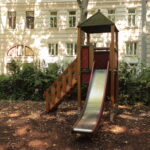 Eine Metallrutsche mit Holztreppe und Unterstand steht in einem schattigen Spielplatzbereich, umgeben von Bäumen und einem weißen Wohnhaus im Hintergrund.